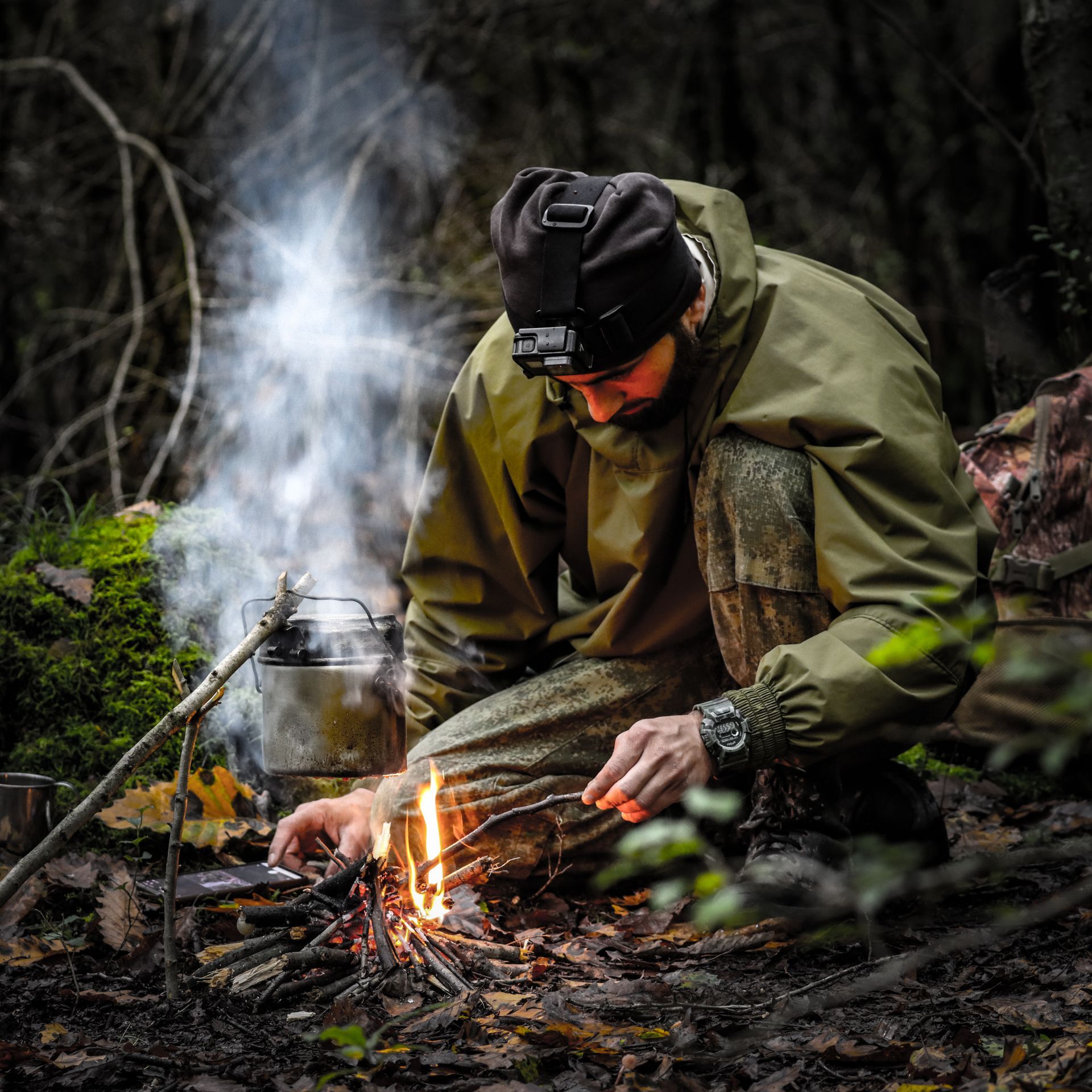 A man in a rain jacket and headlamp feeds sticks to a small campfire, with a pot of water boiling over it in a damp forest.